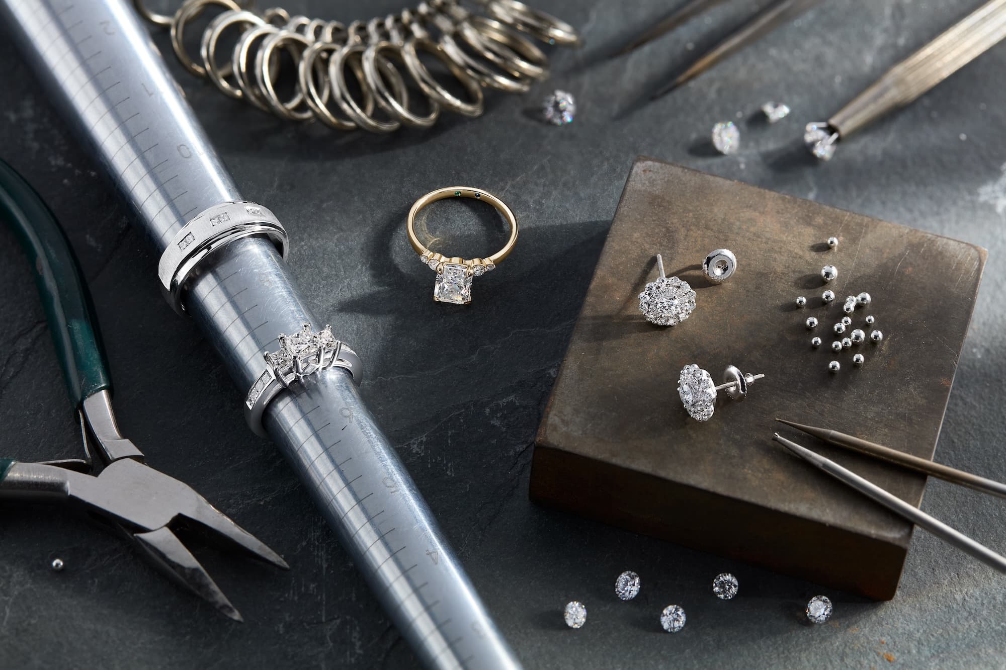 Jewelry repair tools and materials arranged on a dark workbench, including pliers, a ring mandrel with silver rings, a gold engagement ring, loose diamonds, metal beads, tweezers, and a metal block displaying diamond earrings.
