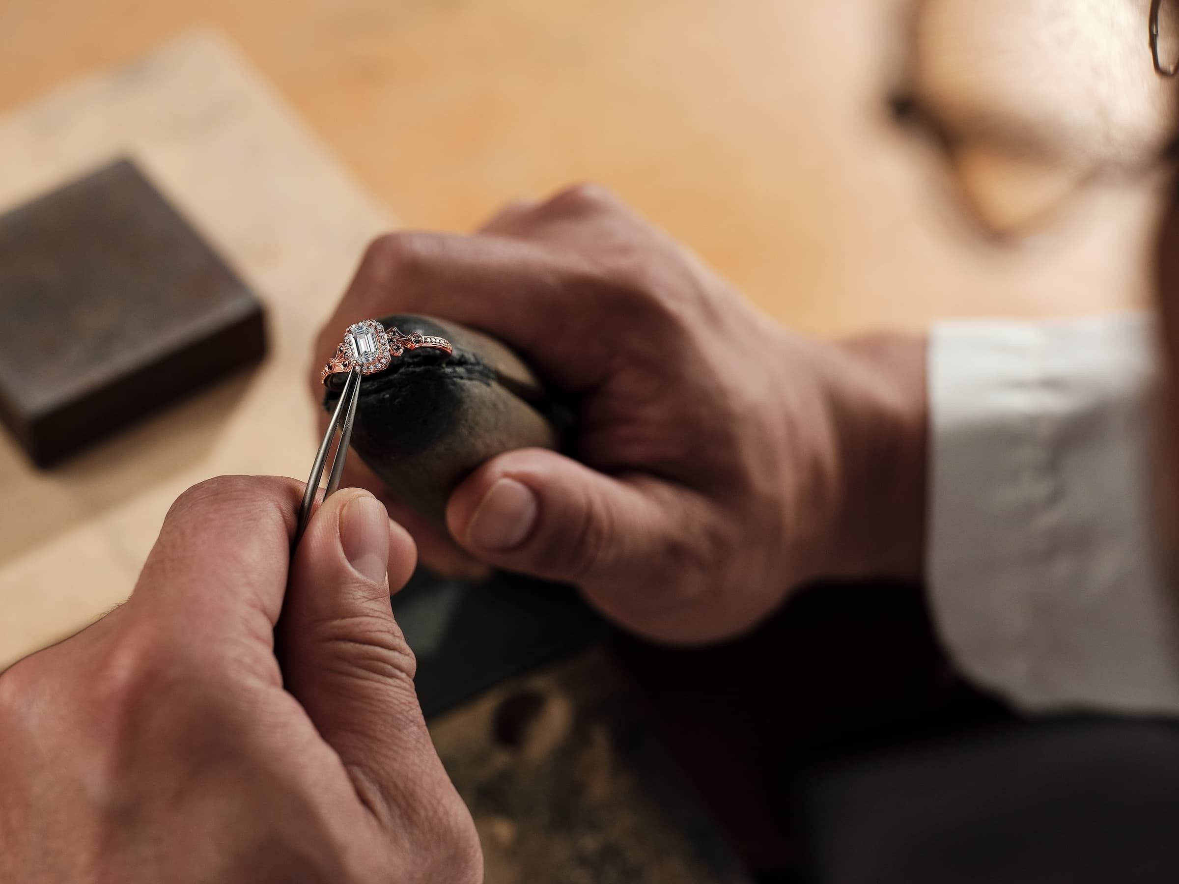 Close-up of hands working on a diamond ring with tools on a wooden surface.